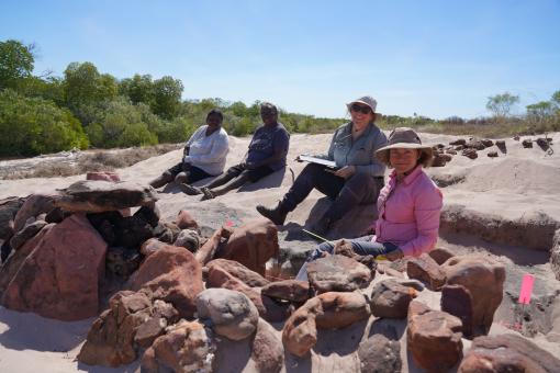 A group of 4 people sitting in a rocky landscape beneath a blue sky